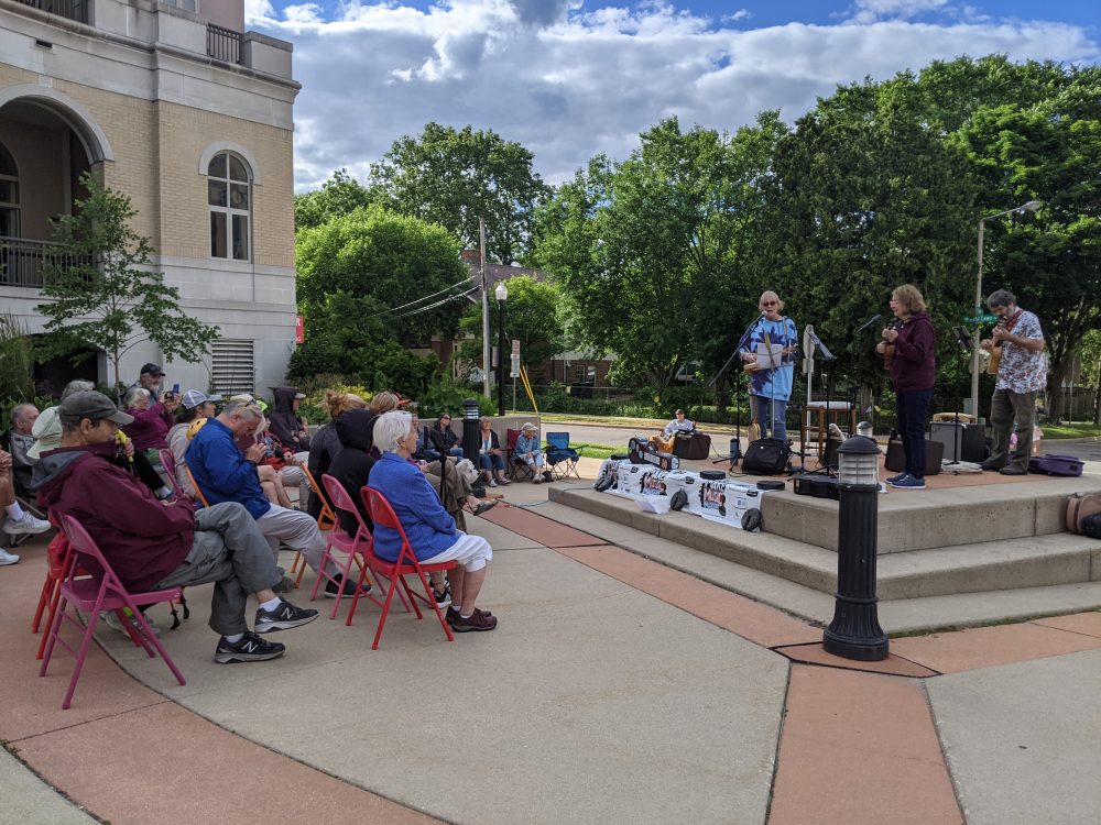 A scene of seated people surrounding an outdoor concrete plaza with three performers playing stringed instruments