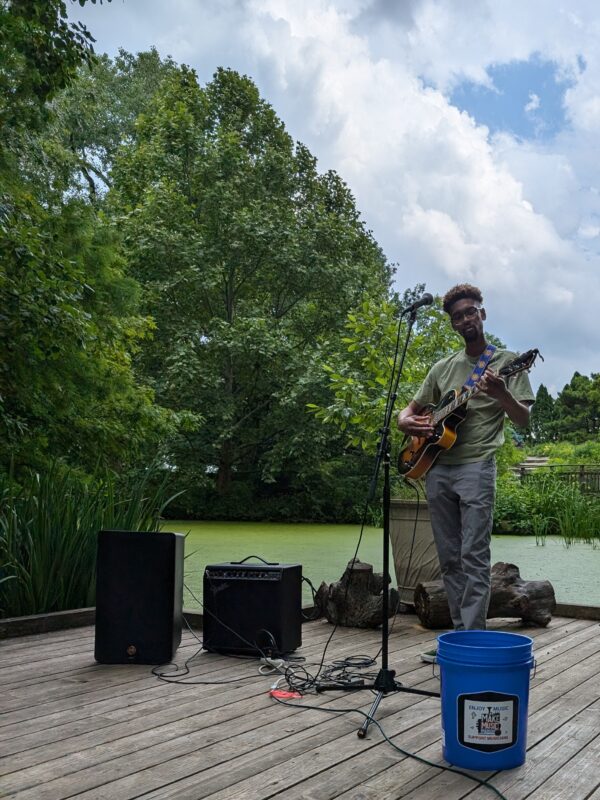 A guitarist plays outside on a porch in front of a pond