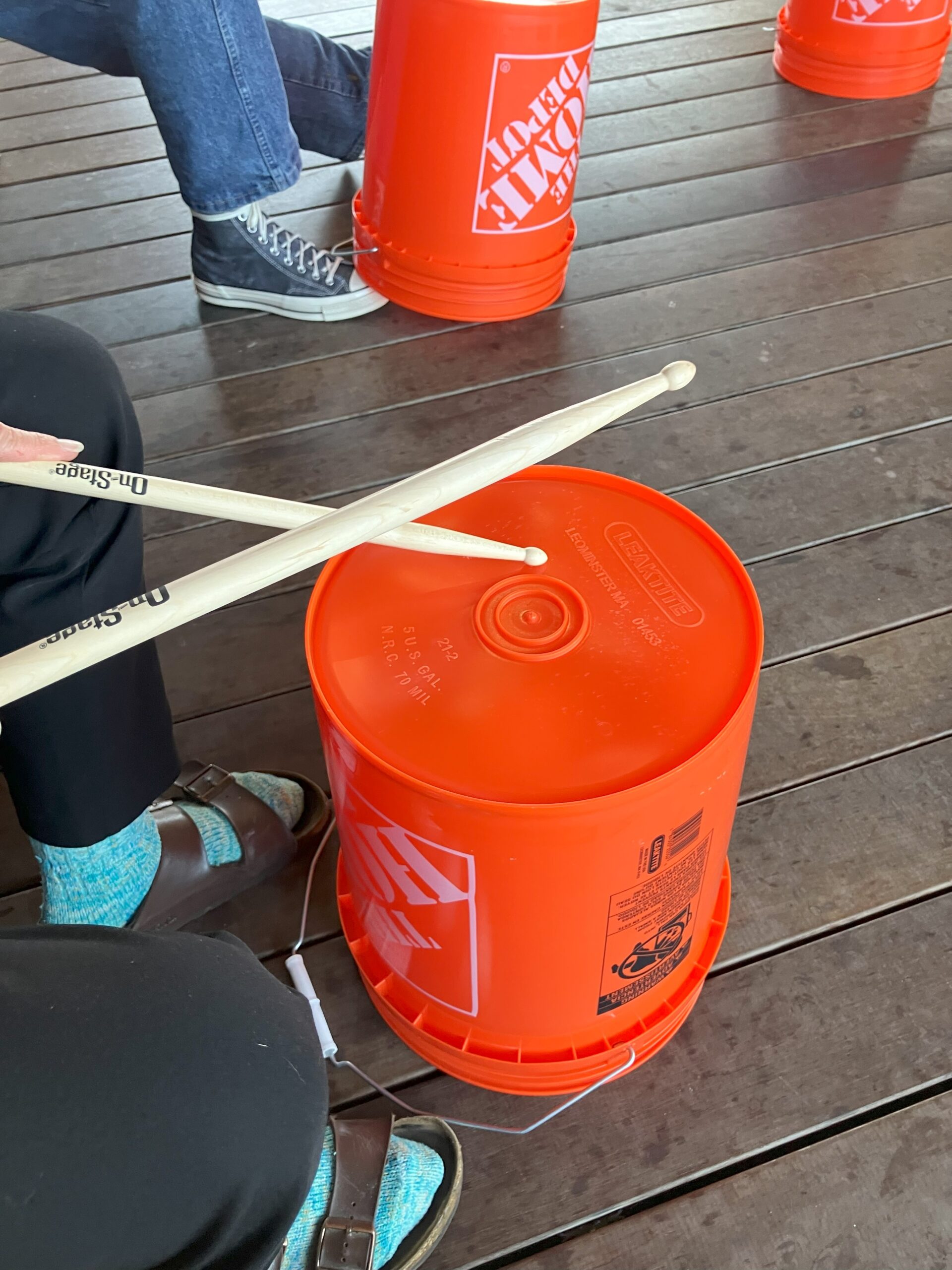 Orange buckets upside down on exterior flooring with participant legs and drum sticks in the corner.