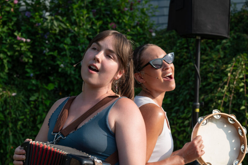 Two women singing. One while playing the accordion and the other while playing the tambourine.