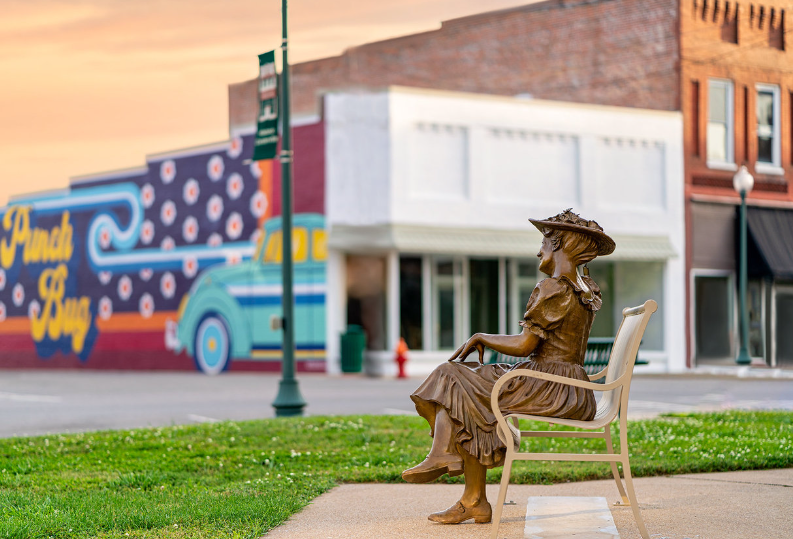 Sunset photograph of the Minnie Pearl statue sitting on a park bench across from a mural with a VW bug in downtown Centerville, TN