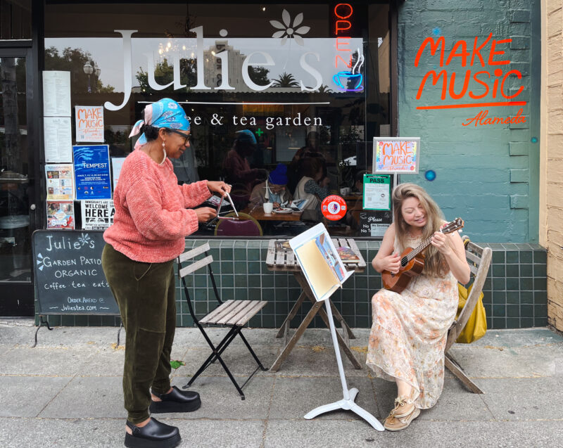 A patron at Julies Coffee and Tea garden plays the triangle with Make Music Alameda 2026 organizer Katherine Park as she sings and plays ukulele on the sidewalk. Photo by Douglas Despres