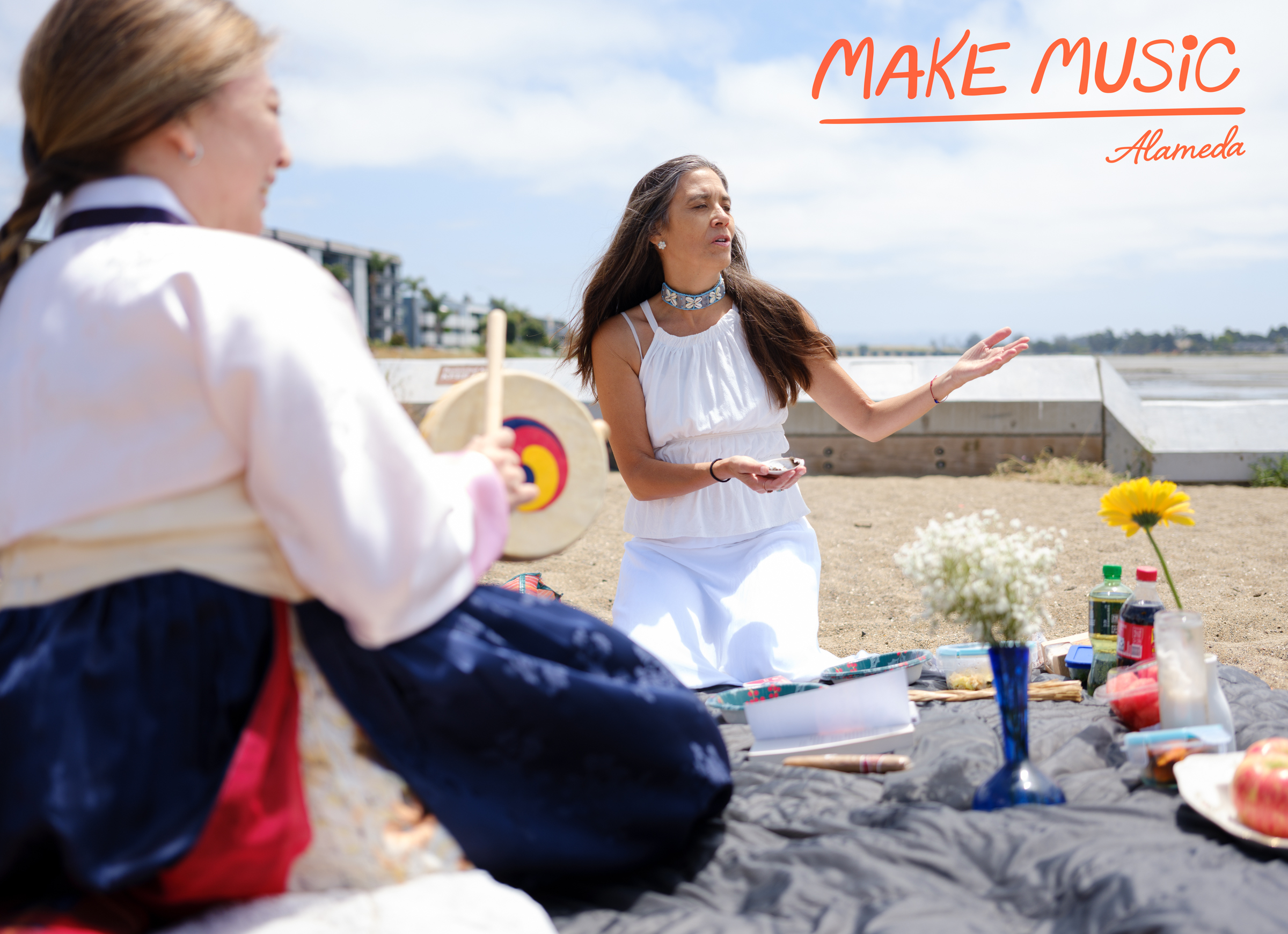 Katherine Park dressed in hanbok, strikes the sogo drum while Alison Hart, wearing a shell necklace and dressed in white sings, they sit on a blanket on the beach with offerings of white flowers, food and drinks for acknowledging their ancestors. Photo by Douglas Despres