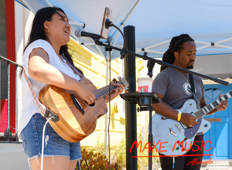a woman sings into a microphone while strumming an acoustic guitar, a man strums an electric guitar