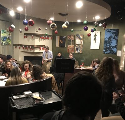 Trio of musicians in a crowded bar decorated for the holidays. A male vocalist performs in front of a guitar player and percussionist on a cajon